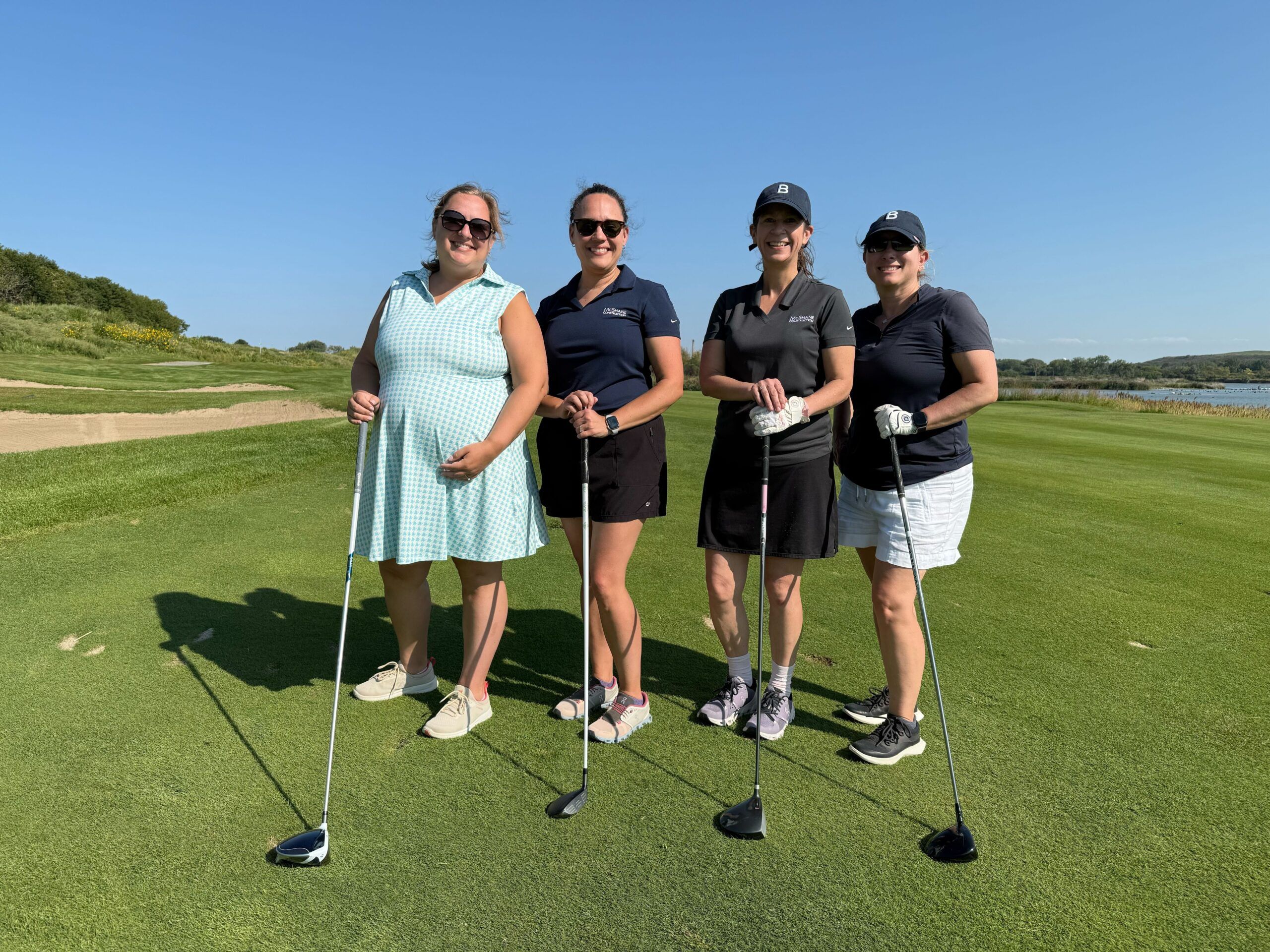 Four women stand on a golf course, holding clubs and smiling at the camera.